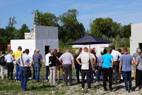 Gruppe von Menschen steht vor zwei weißen Testgebäuden bei einer Präsentation.