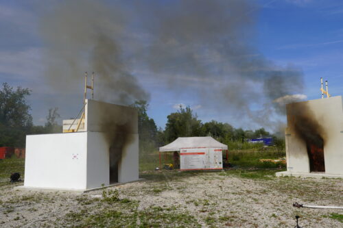 Zwei Testgebäude, beide mit Rauchentwicklung, eines bereits in Flammen.
