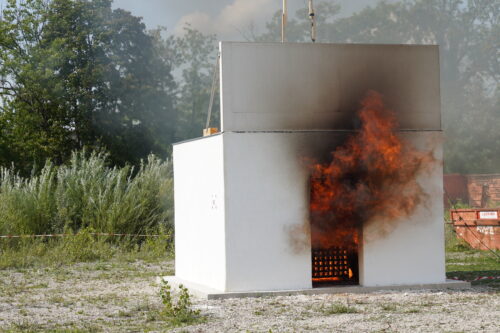 Ein weißes Testgebäude, aus dessen Türöffnung Flammen und schwarzer Rauch schlagen