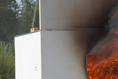 Versuchshaus mit weißer Fassade, dessen Innenraum in Flammen steht; starke Feuerentwicklung schlägt aus der geöffneten Tür, während die obere Hälfte des Gebäudes schwarz vom Rauch verfärbt ist. Im Hintergrund sind Bäume.