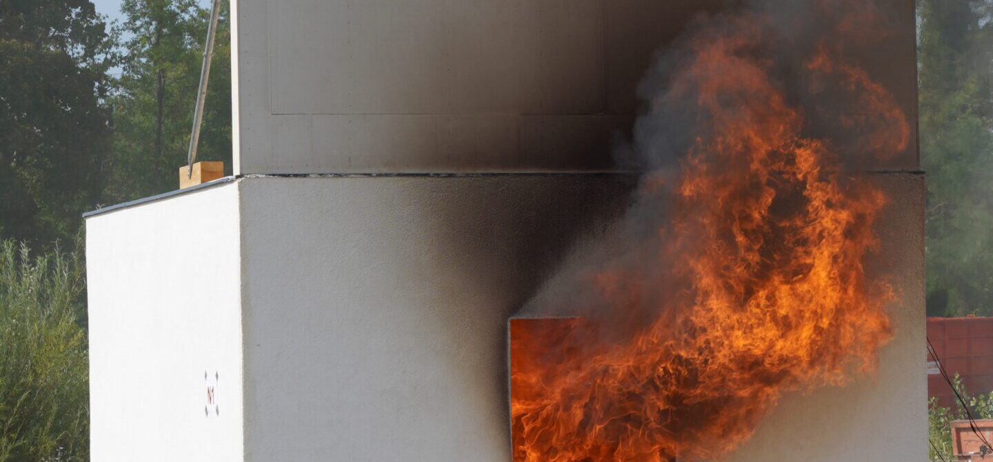 Versuchshaus mit weißer Fassade, dessen Innenraum in Flammen steht; starke Feuerentwicklung schlägt aus der geöffneten Tür, während die obere Hälfte des Gebäudes schwarz vom Rauch verfärbt ist. Im Hintergrund sind Bäume.