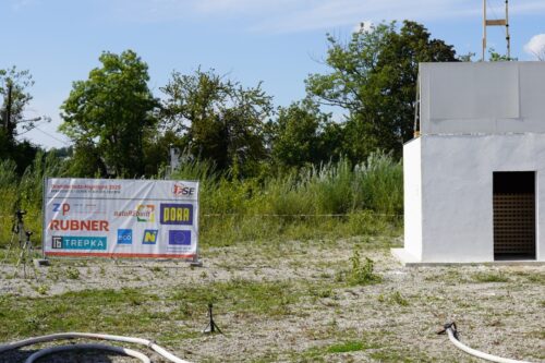 Weißes Testgebäude mit offener Tür, daneben ein Banner mit Sponsorenlogos.