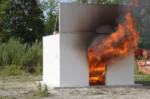 Ein weißes Testgebäude, aus dessen Türöffnung Flammen und schwarzer Rauch schlagen.