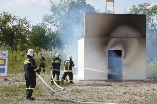 Feuerwehrleute löschen ein stark verrußtes Testgebäude mit Wasserstrahlen.