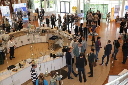 Menschen stehen in kleinen Gruppen in einem hellen Veranstaltungsraum zusammen. In der Mitte befindet sich eine gebogene Theke mit Kaffee und Getränken. Die Szene zeigt eine Networking-Situation während einer Messe oder Tagung.