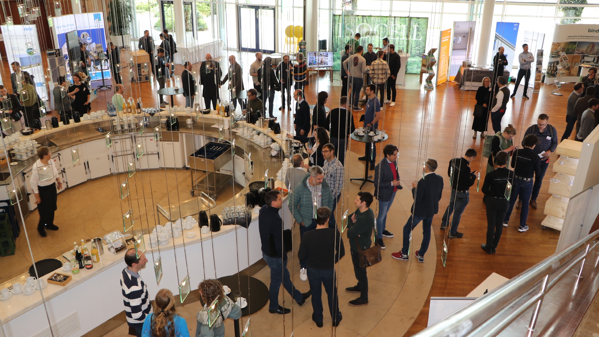 Menschen stehen in kleinen Gruppen in einem hellen Veranstaltungsraum zusammen. In der Mitte befindet sich eine gebogene Theke mit Kaffee und Getränken. Die Szene zeigt eine Networking-Situation während einer Messe oder Tagung.