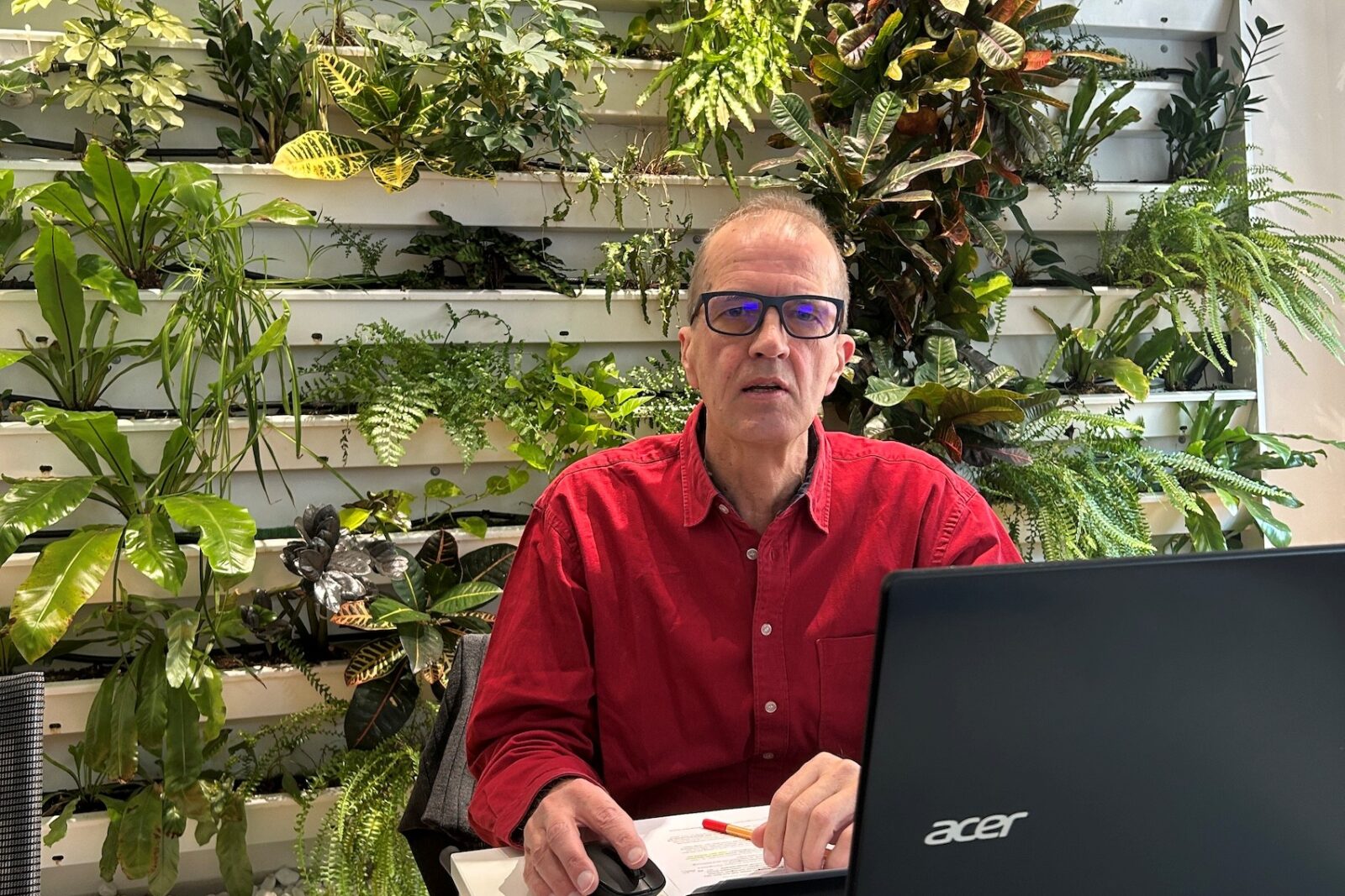 Ein Mann mit rotem Hemd und Brille sitzt an einem Schreibtisch vor einem Laptop. Hinter ihm befindet sich eine große vertikale Pflanzenwand mit verschiedenen Grünpflanzen. Auf dem Tisch liegen ein Smartphone, Unterlagen, ein Stift und ein Glas Wasser.