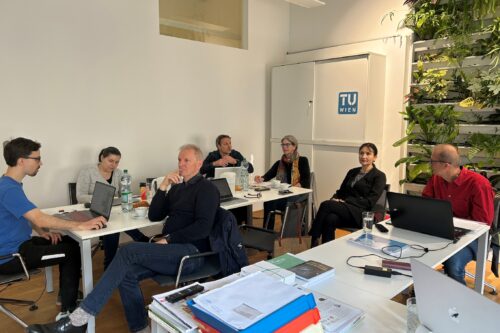 Mehrere Personen sitzen in einem hellen Besprechungsraum an Tischen mit Laptops und Notizen. An der Wand ist ein „TU Wien“-Logo zu sehen, rechts daneben eine bepflanzte Wand. Die Gruppe wirkt konzentriert und befindet sich in einer Besprechung oder Präsentation.