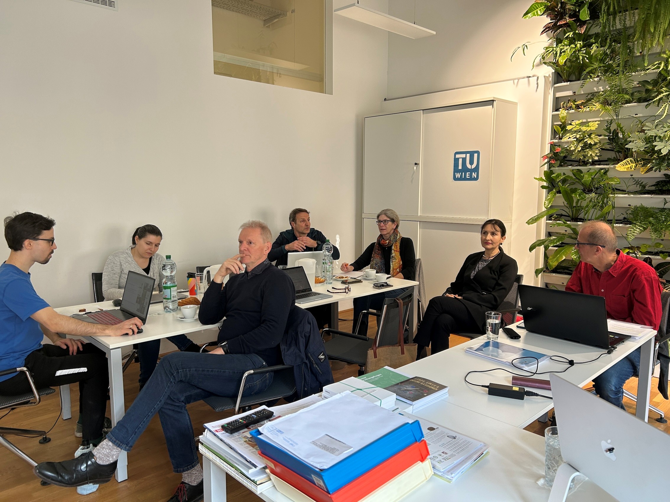 Mehrere Personen sitzen in einem hellen Besprechungsraum an Tischen mit Laptops und Notizen. An der Wand ist ein „TU Wien“-Logo zu sehen, rechts daneben eine bepflanzte Wand. Die Gruppe wirkt konzentriert und befindet sich in einer Besprechung oder Präsentation.