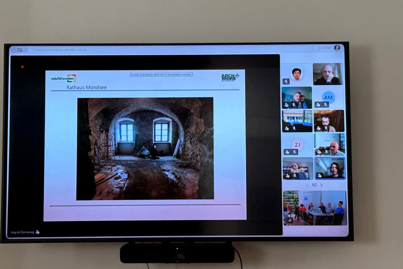 Ein großer Bildschirm zeigt eine Online-Präsentation mit dem Titel „Rathaus Mondsee“ und einem Foto eines gewölbten, renovierungsbedürftigen Raums. Rechts sind mehrere Videokonferenz-Teilnehmende in kleinen Fenstern zu sehen.